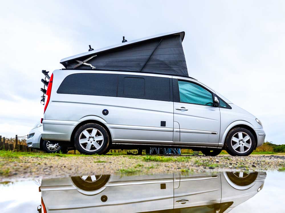 minivan-conversion A converted minivan with a pop-up top sits in front of a puddle, a reflection of the minivan shows in the puddle in the foreground