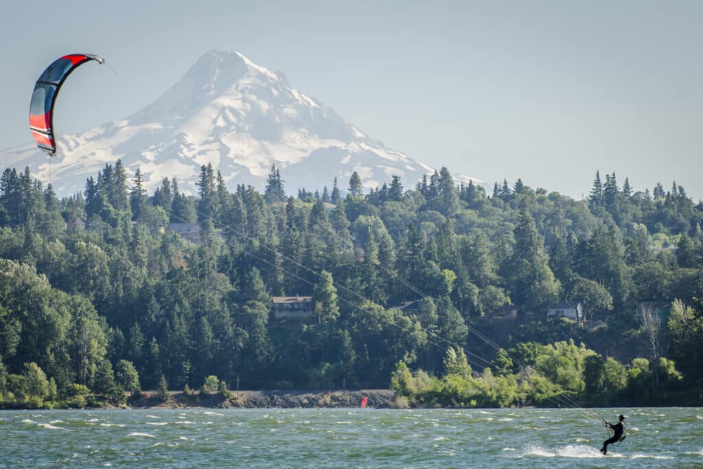 Landscape of Mount Hood in Oregon USA green pine trees in front of the huge snowy mountain with one person kiteboarding on the Columbia river in the foreground. from Tumbleweed Travel